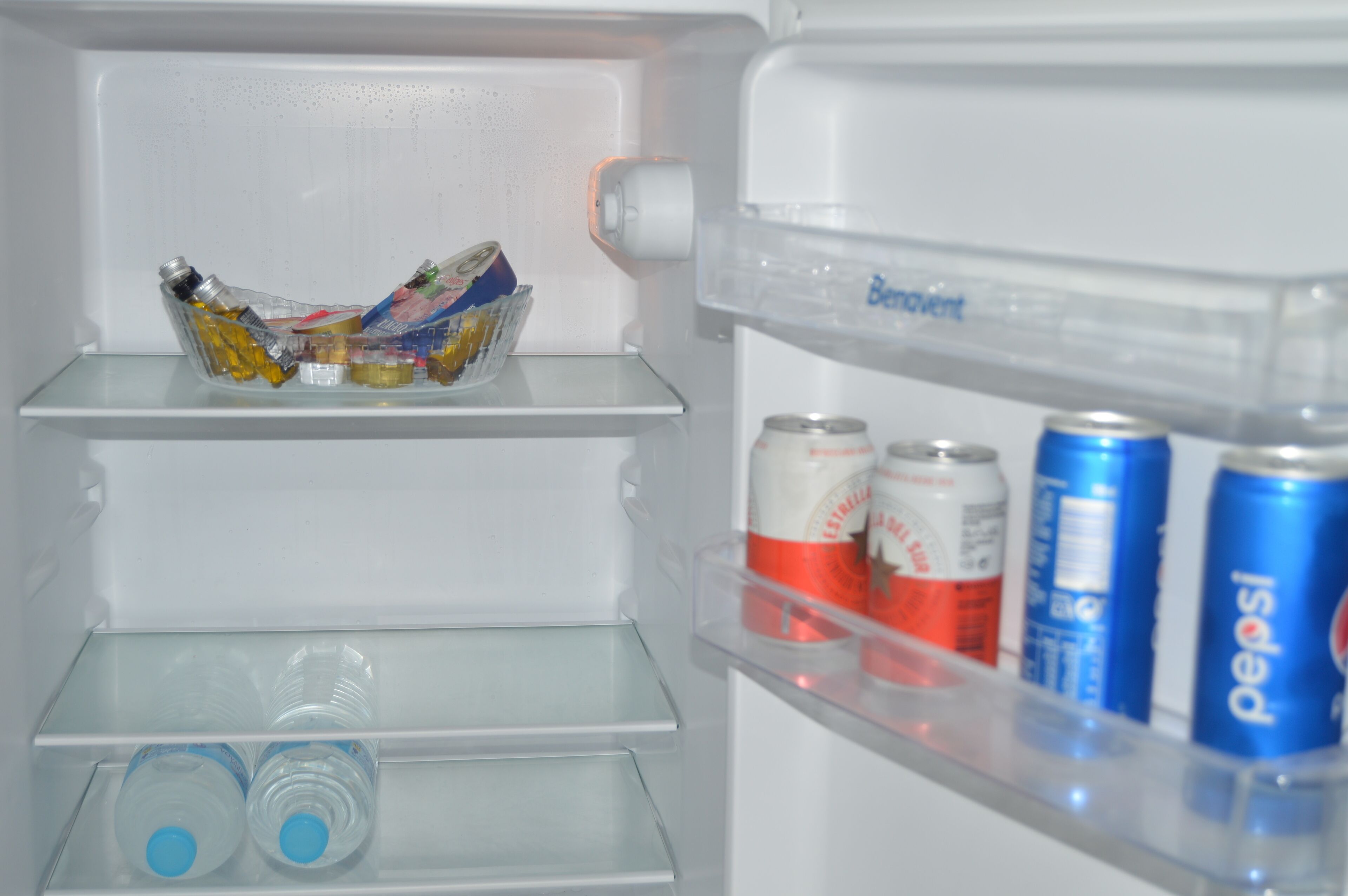 Un refrigerador amb una prateleira transparent, un bol de vidre amb condiments, i tres llaunes de Pepsi.