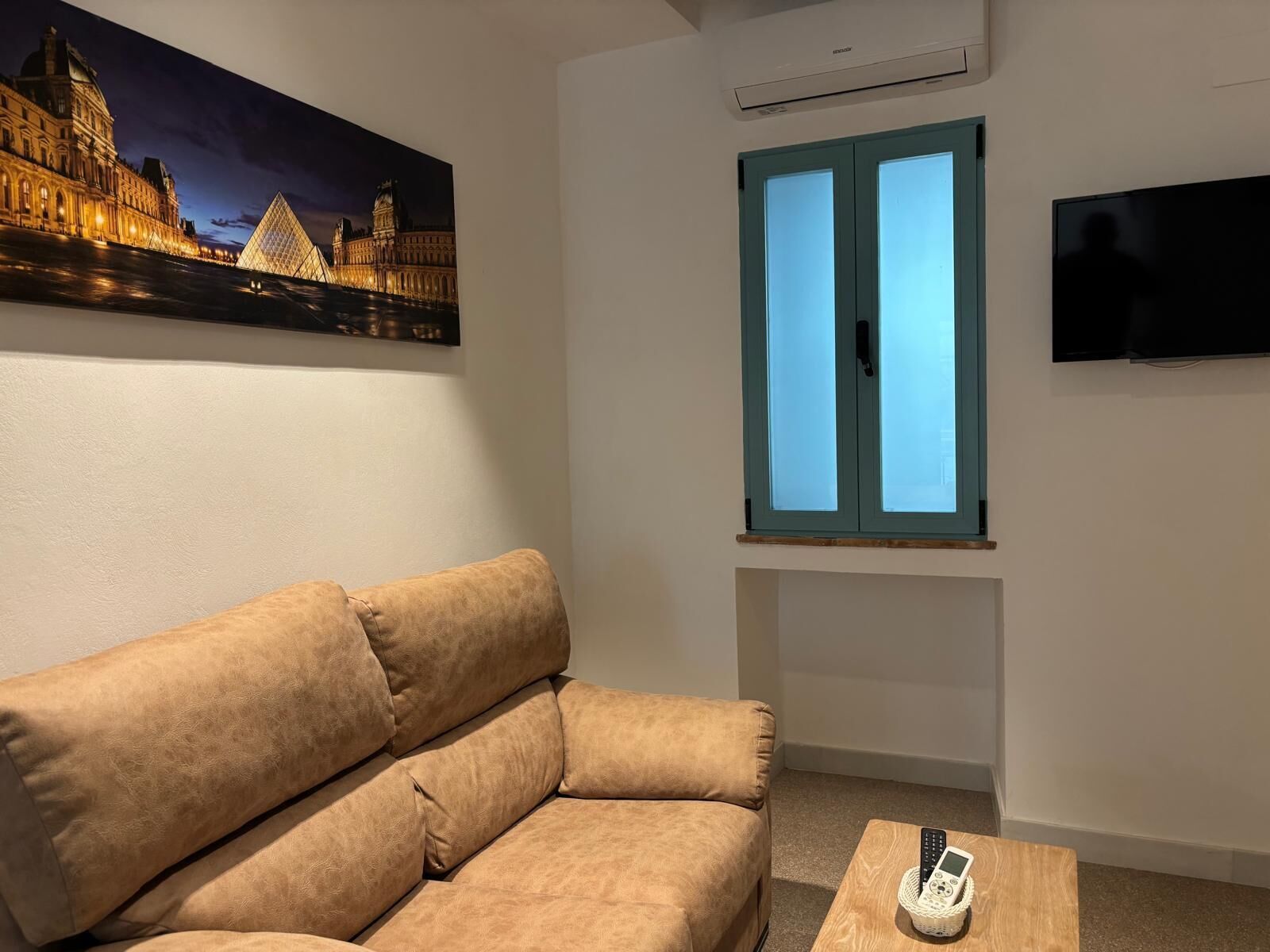 Una sala de estar con un sofá, una televisión, una ventana con vista a un edificio y un equipo de aire acondicionado.