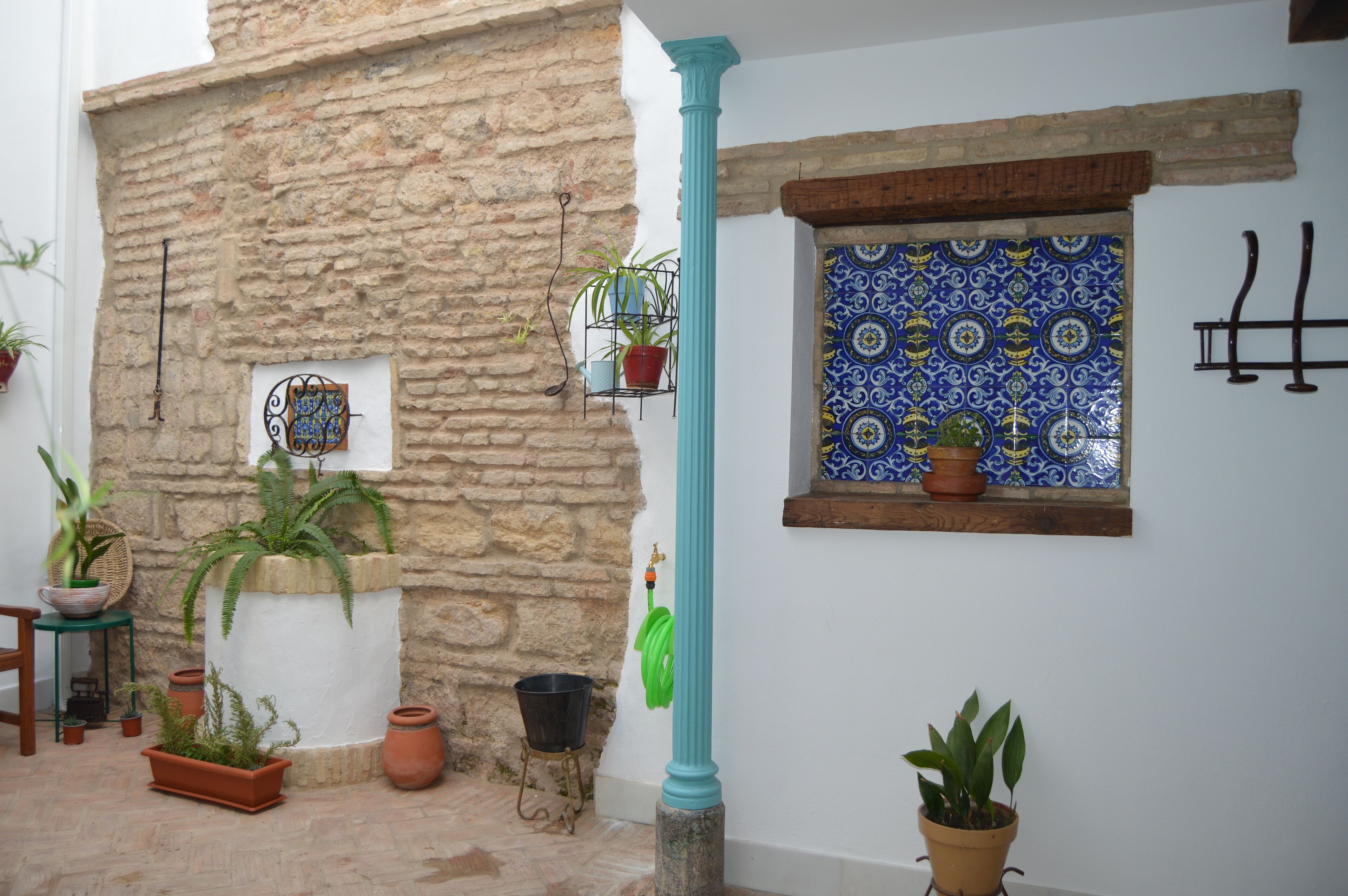 Un mur de pierre agrémenté de plantes en pot, une fenêtre ornée de carreaux bleus et un tuyau d’arrosage vert suspendu.