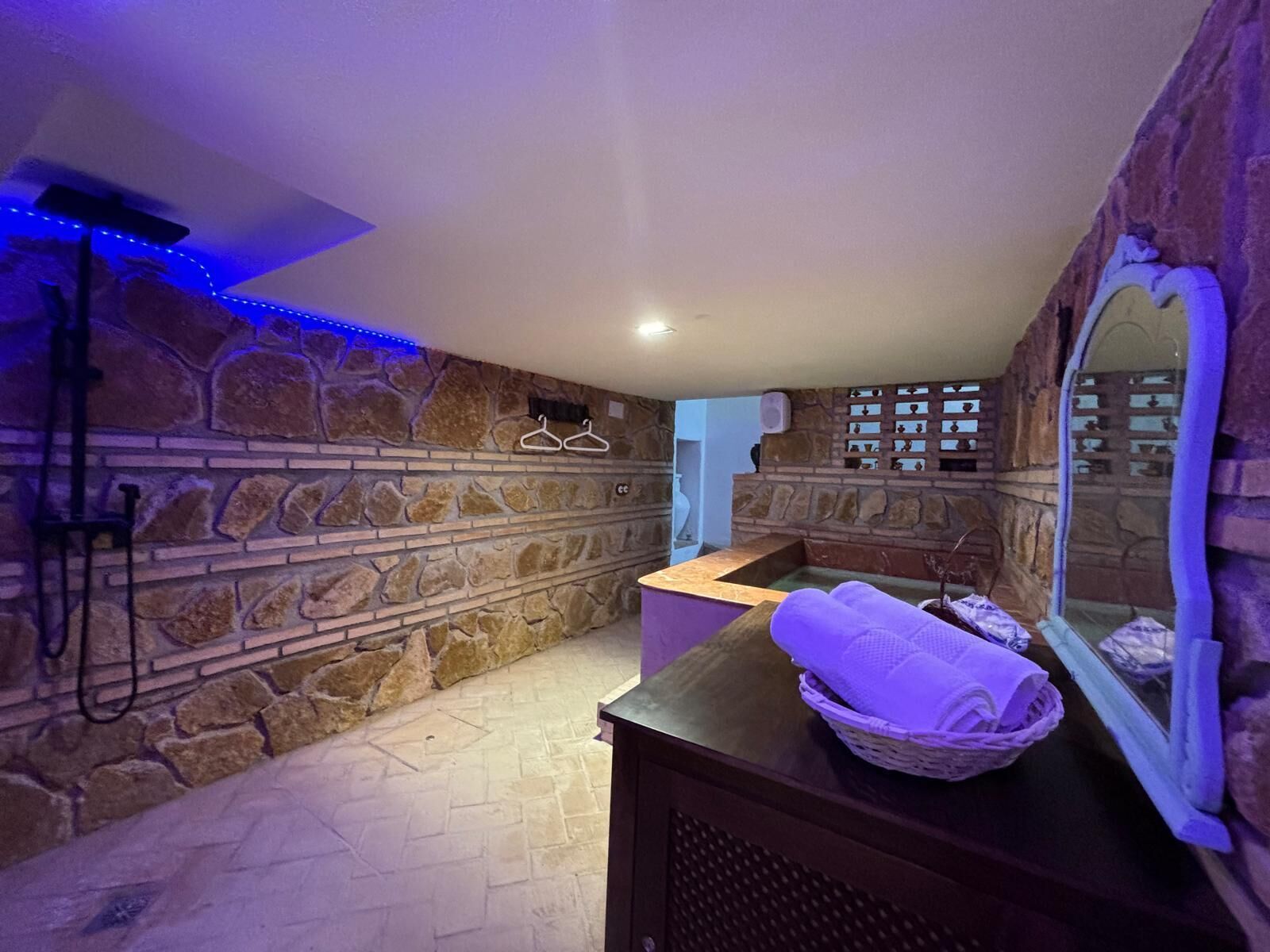 Un baño con paredes y suelo de piedra, un tocador de madera con espejo, y una cesta de toallas.
