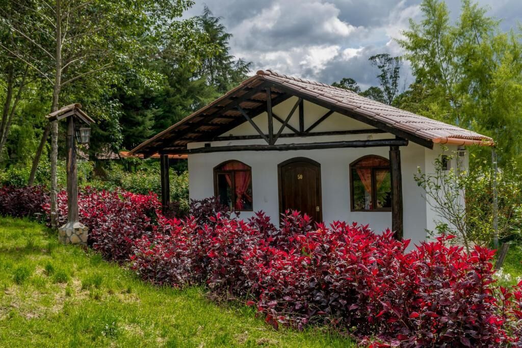Un edificio de estilo chalete tradicional con techo inclinado y vigas de madera, rodeado de abundante follaje y plantas de hojas rojas.