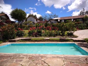 Ein Schwimmbecken mit Steinplatten, umgeben von einem Garten mit verschiedenen Pflanzen und Blumen, und Häusern im Hintergrund.