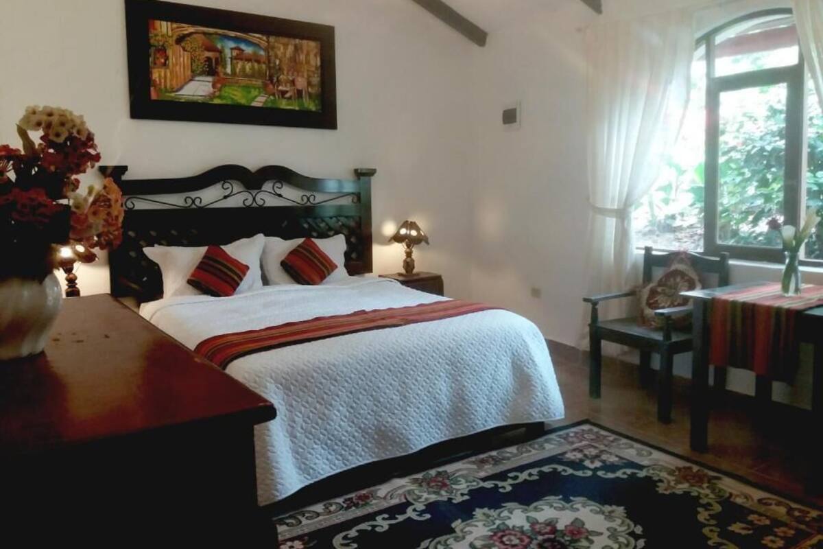 A bedroom with a bed, a nightstand, a chair, a table, a painting, and a window.