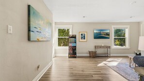 A spacious room with hardwood flooring, a large window, a painting, a gaming console, and a chair.