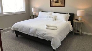 A neatly made bed with white linens and pillows, two bedside tables with lamps, a framed picture on the wall, and a window with blinds. - Newly UPD home in DT Libertyville Near Naval Base! (Libertyville)