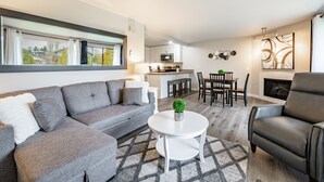 Ein modernes Wohnzimmer mit einem grauen Sofa, einem schwarzen Ledersessel, einem weißen Couchtisch und einem Teppich mit Muster. Im Hintergrund ist eine Küchenzeile mit Esstisch und Stühlen zu sehen.