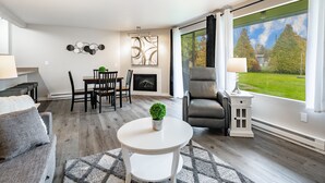Ein modernes Wohnzimmer mit Kamin, Essbereich und großen Fenstern mit Blick auf eine grüne Landschaft.