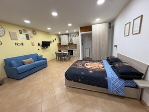 A bedroom with a bed, a blue sofa, a small table, and a kitchenette in the background.