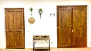 Ein Zimmer mit zwei Holztüren, einem kleinen Tisch mit Schubladen, einem an der Wand befestigten Regal mit Pflanzen und einer Wanduhr.