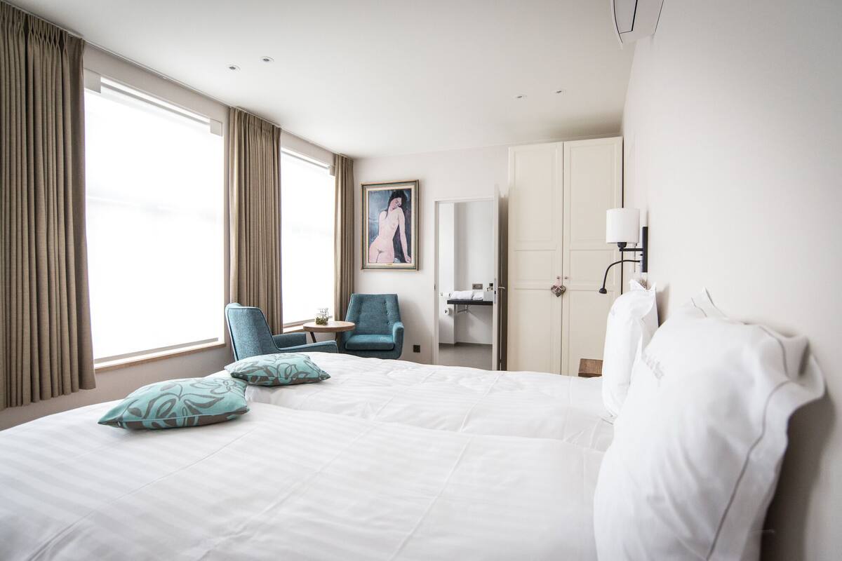 A hotel room with a large bed, two chairs, a desk, a painting, a wardrobe, and a bathroom visible through an open door.