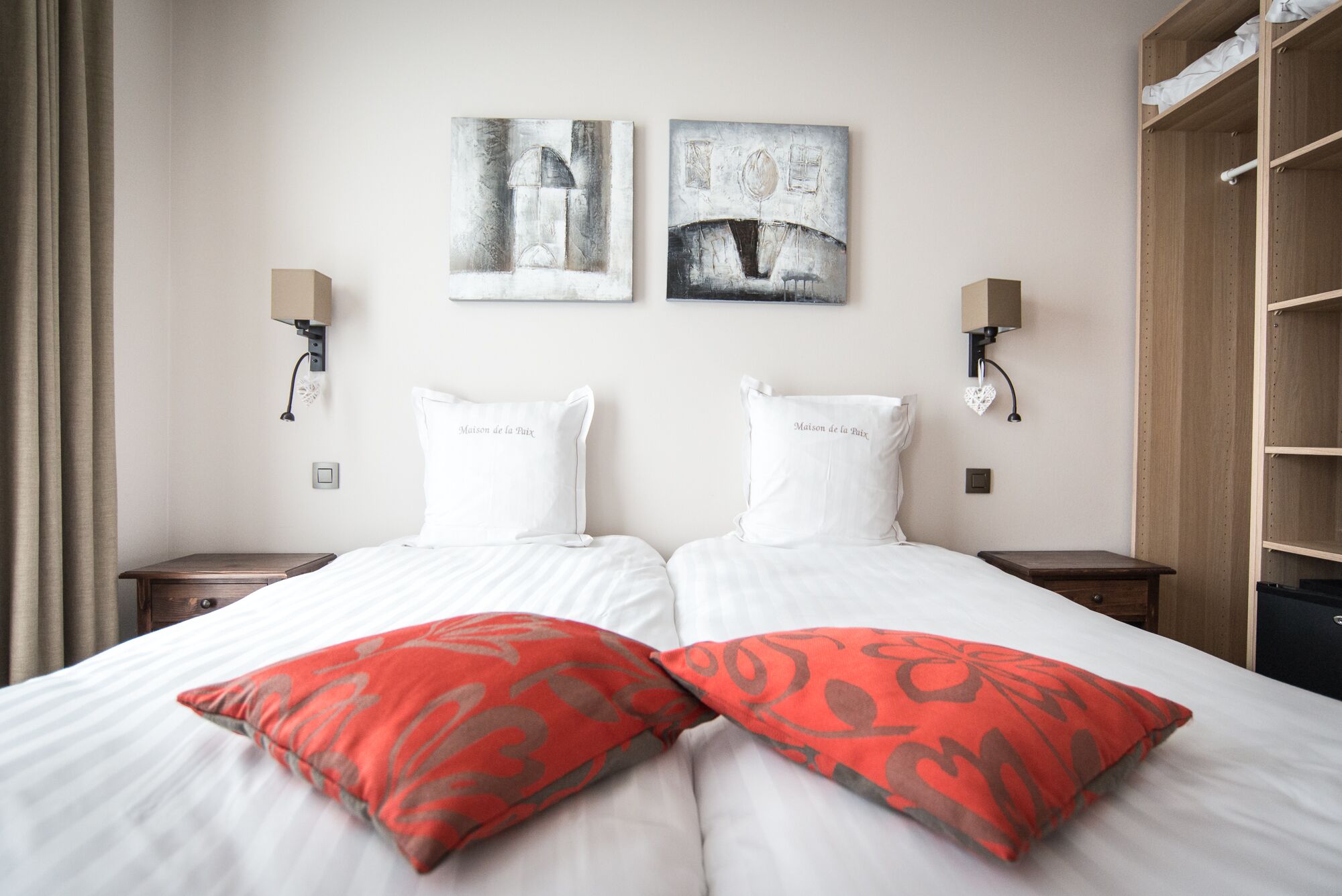 A hotel room with two beds, each with white pillows and a red patterned pillow. There are two wall-mounted lamps on either side of the beds.