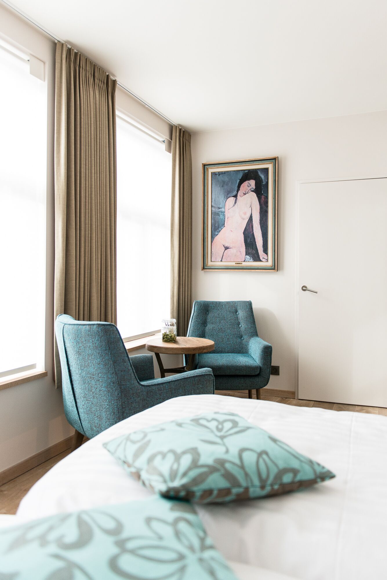 A hotel room with a bed, two chairs, a small table, a painting on the wall, and a window with curtains.