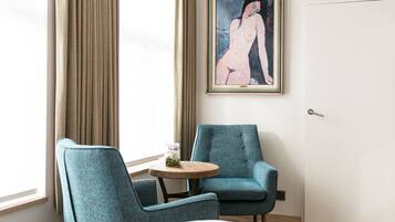 A hotel room with a bed, two chairs, a small table, a painting on the wall, and a window with curtains.