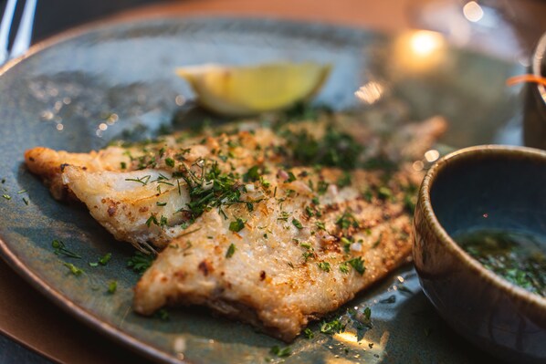 A plate with a piece of fish garnished with herbs and a lemon wedge, accompanied by a bowl of sauce. - Maison de la Paix (Poperinge)