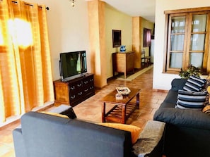 Una sala de estar con un sofá, un televisor sobre un mueble de madera, una mesa de centro y una ventana con cortinas.