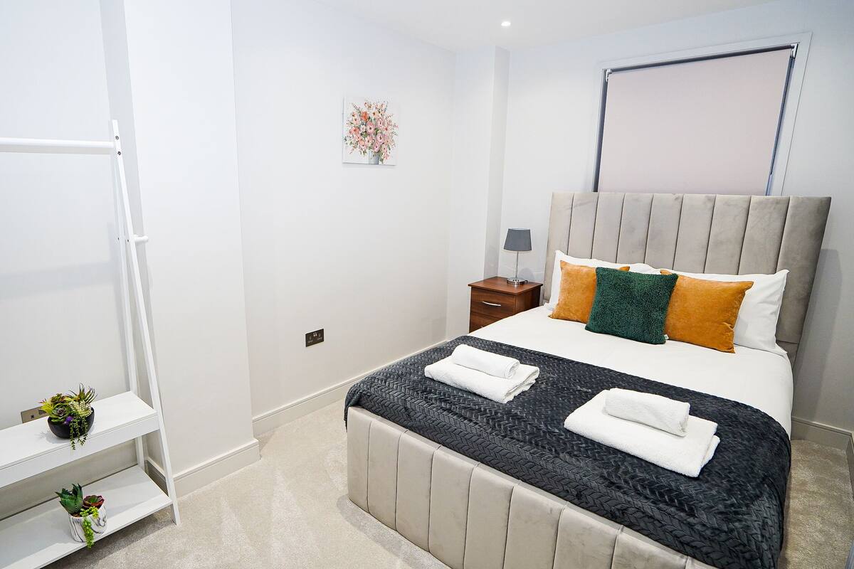 Quarto com cama, mesa de cabeceira, abajur e prateleira na parede com plantas.