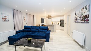 A modern living room with a blue sectional sofa, a glass coffee table, and a kitchen in the background. - Penthouses in Slough (Slough)