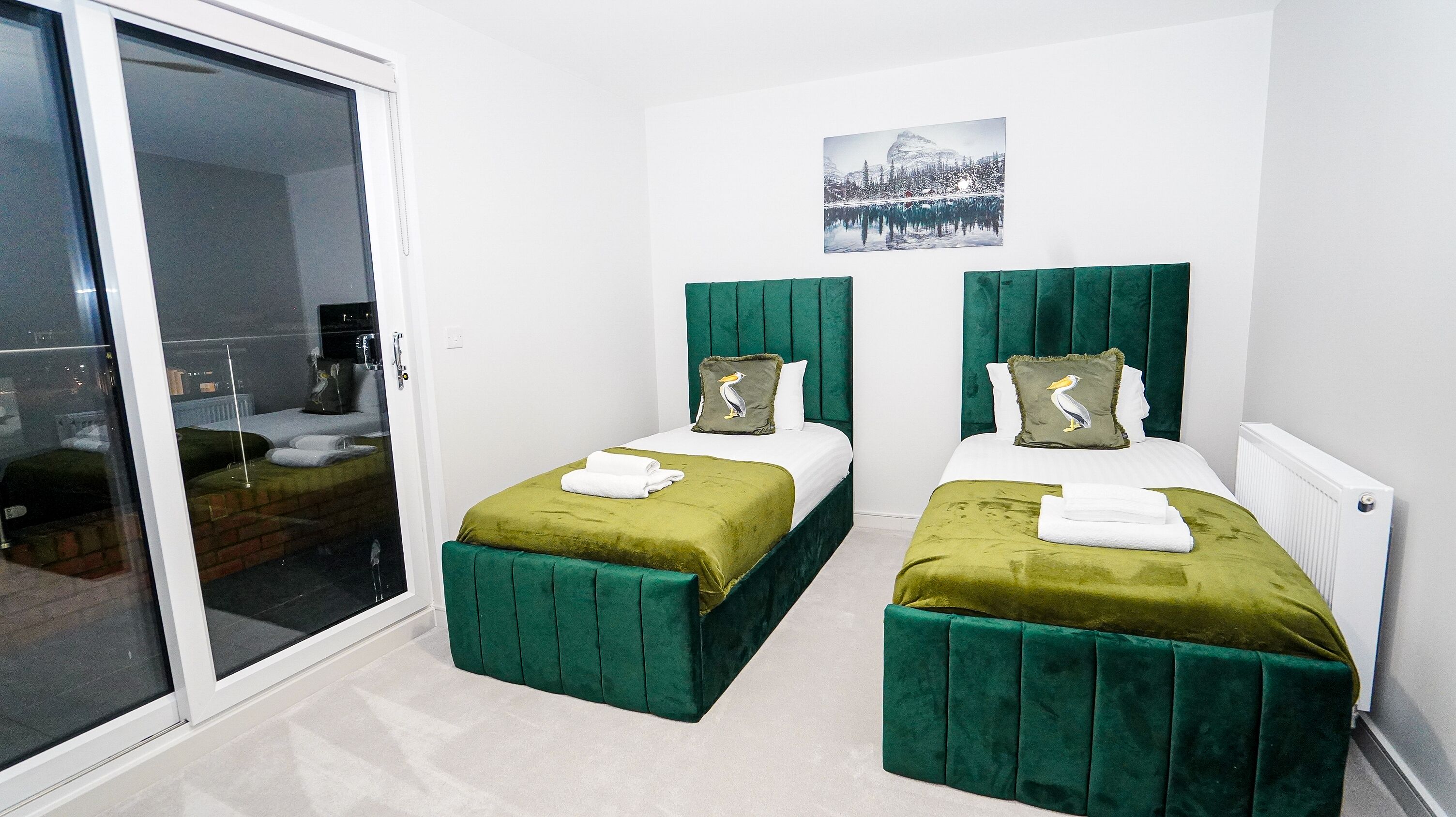 A hotel room with two beds, each with green headboards and matching green footboards. There is a white wall with a framed picture above the beds.