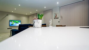 A modern living room with a white marble coffee table, a potted plant, a flat-screen TV, a blue sofa, and a dining area with chairs. - Penthouses in Slough (Slough)