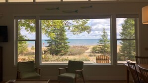 Ein Zimmer mit großem Fenster mit Blick auf Strand und Meer, zwei grüne Stühle, ein Holztisch und ein Deckenventilator.