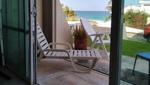 A balcony with white lounge chairs and a potted plant, overlooking a beach and ocean. - Ocean Front Roomy Caribbean Condo (Cancún)