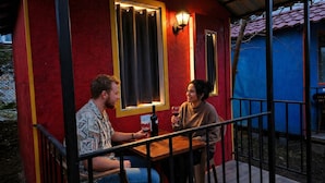 Dos personas sentadas en una mesa al aire libre con baranda, bajo una estructura con paredes rojas y fachada azul.
