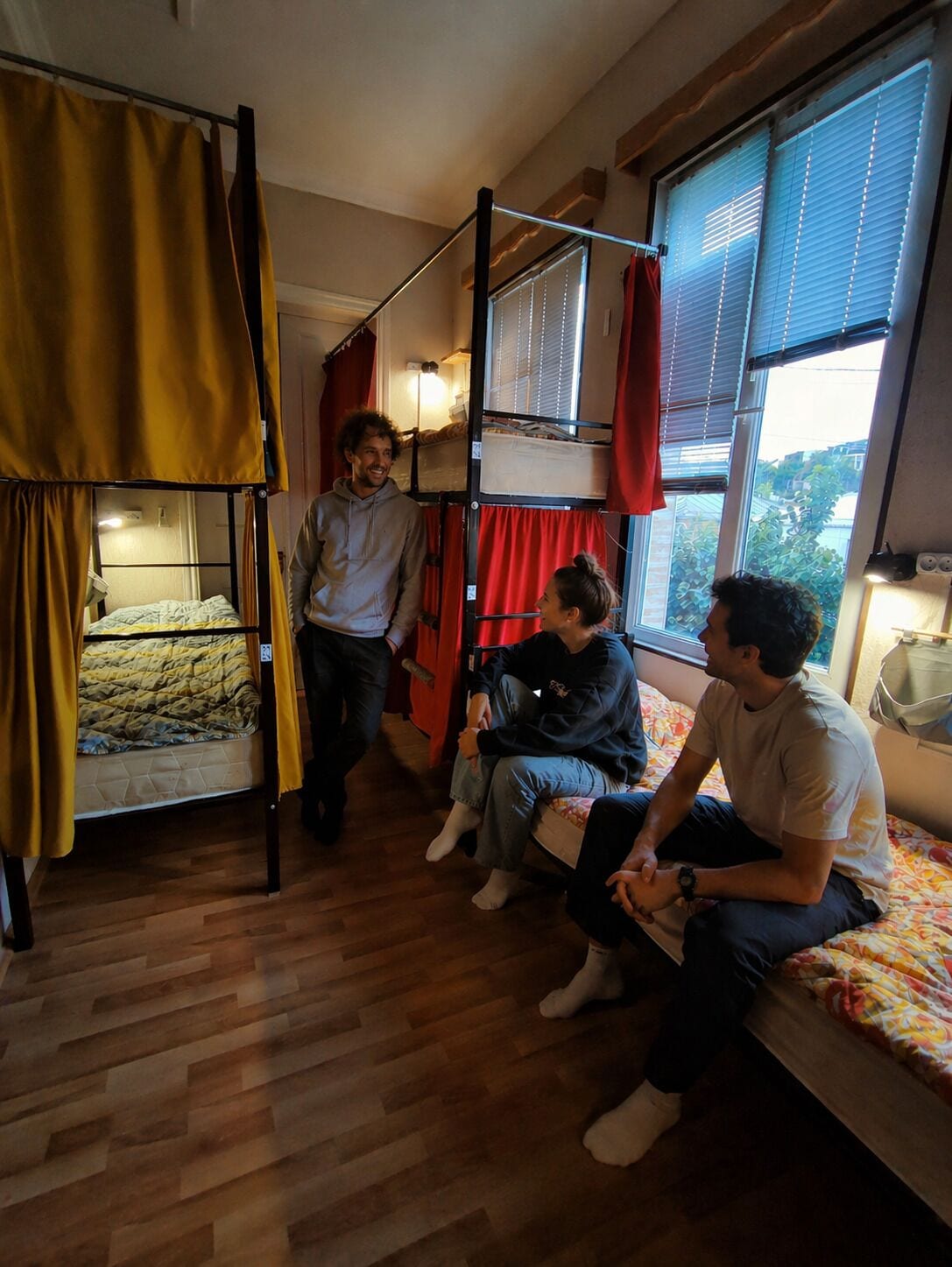 A dormitory room with bunk beds, a wooden floor, and a window with blinds.