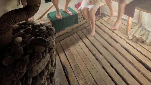 Una sauna con tres personas sentadas en bancos, suelo de madera y un montón de piedras.