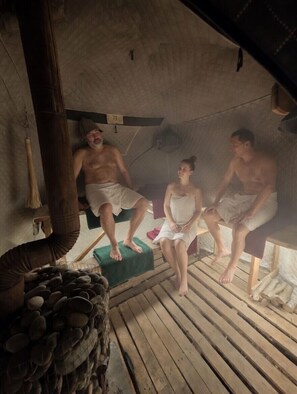 A sauna with three people seated on benches, a wooden floor, and a pile of stones. - The Friends Hostel & Sauna (Kutaisi)
