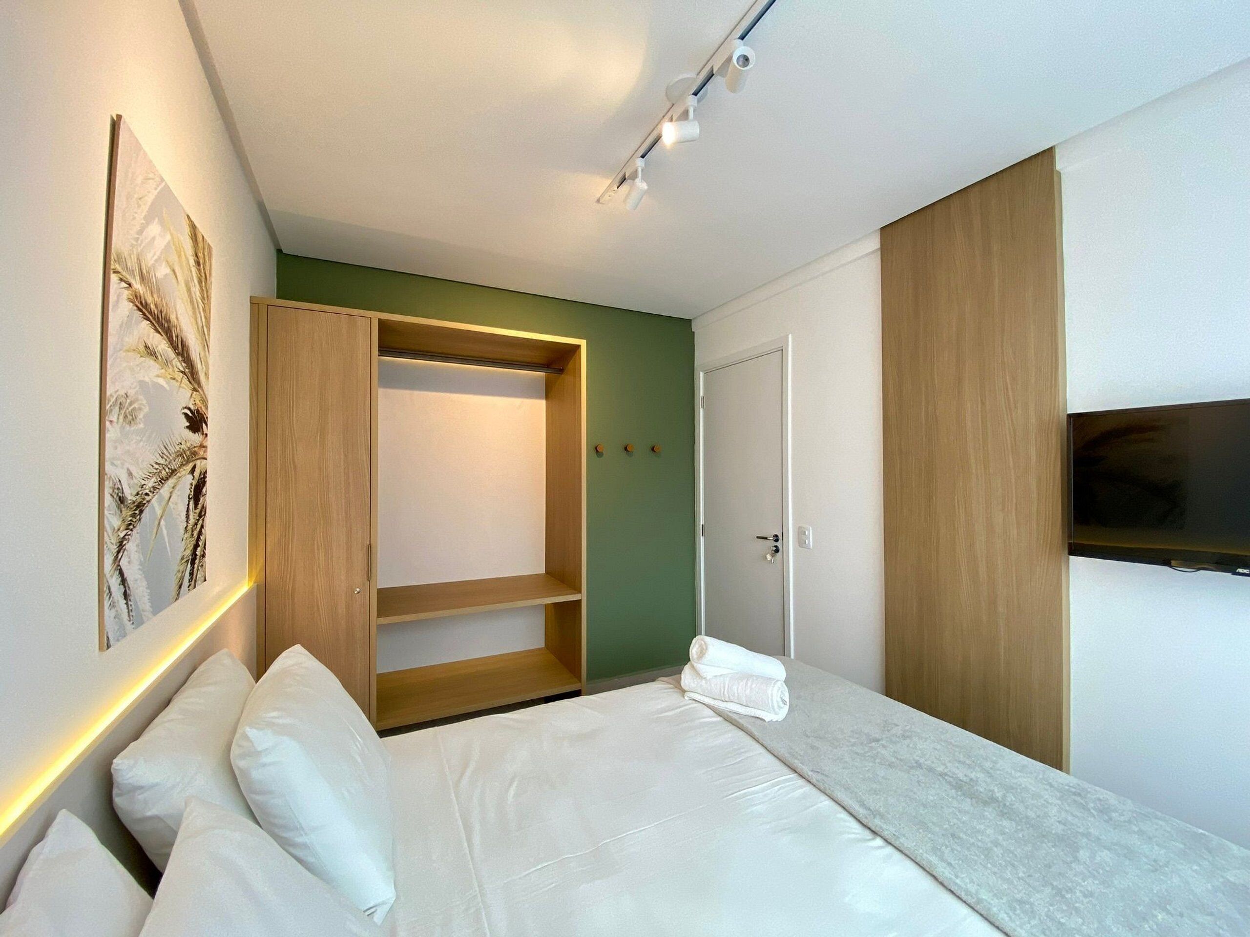 A modern bedroom with a bed, a wooden wardrobe, a green accent wall, and a flat-screen TV.