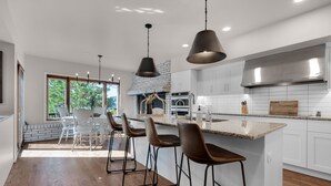 Una cocina moderna con armarios blancos, una isla grande con taburetes de bar, horno empotrado y chimenea de ladrillo.