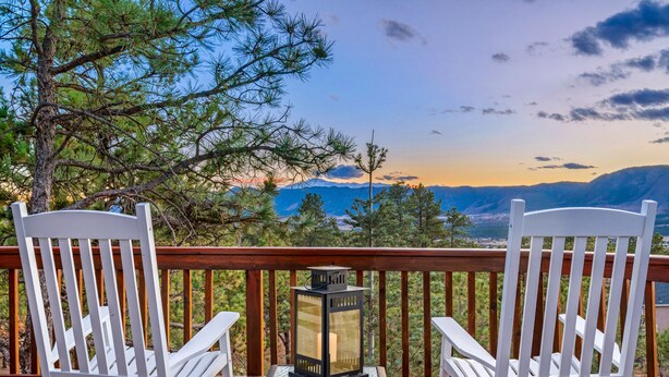 Dos sillas Adirondack blancas sobre una terraza de madera con una mesita de farol, con vistas a un paisaje montañoso.