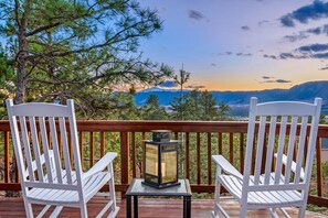 To hvide Adirondack-stole på en træterrasse med et lanternelys-bord, med udsigt over et bjergrigt landskab.