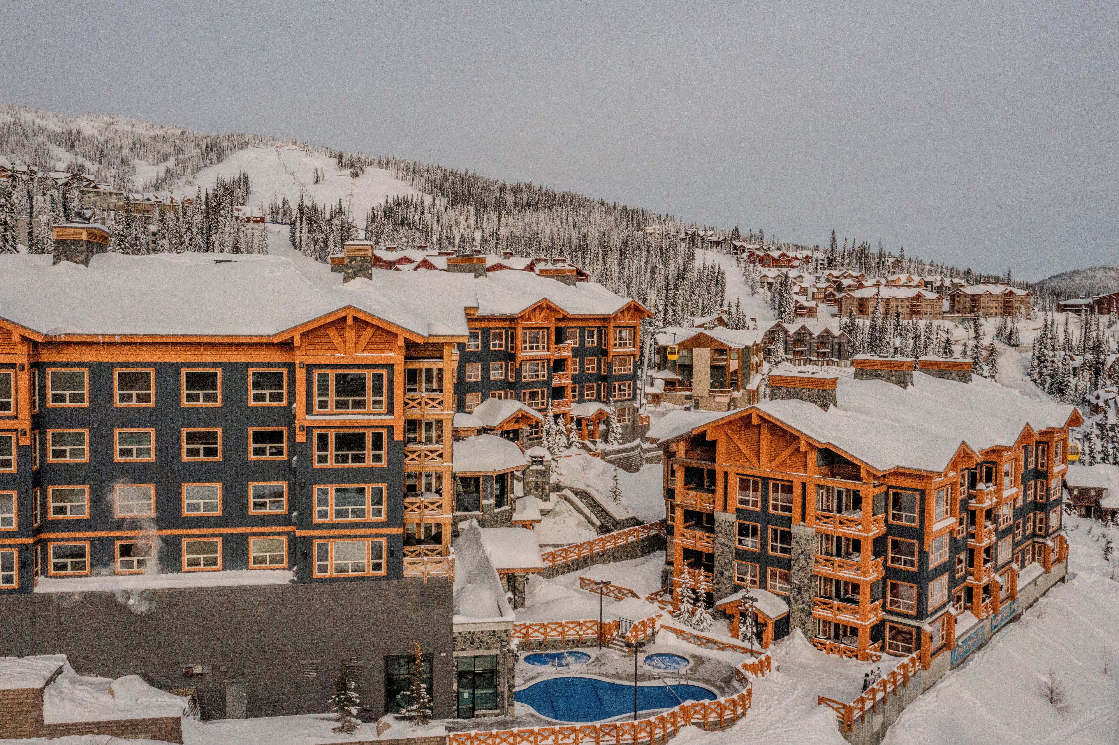 A snow-covered resort with multiple buildings, a pool, and a ski slope in the background.