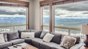 Een moderne woonkamer met een grote hoekbank, twee ramen met zonwering en uitzicht op een besneeerd landschap.