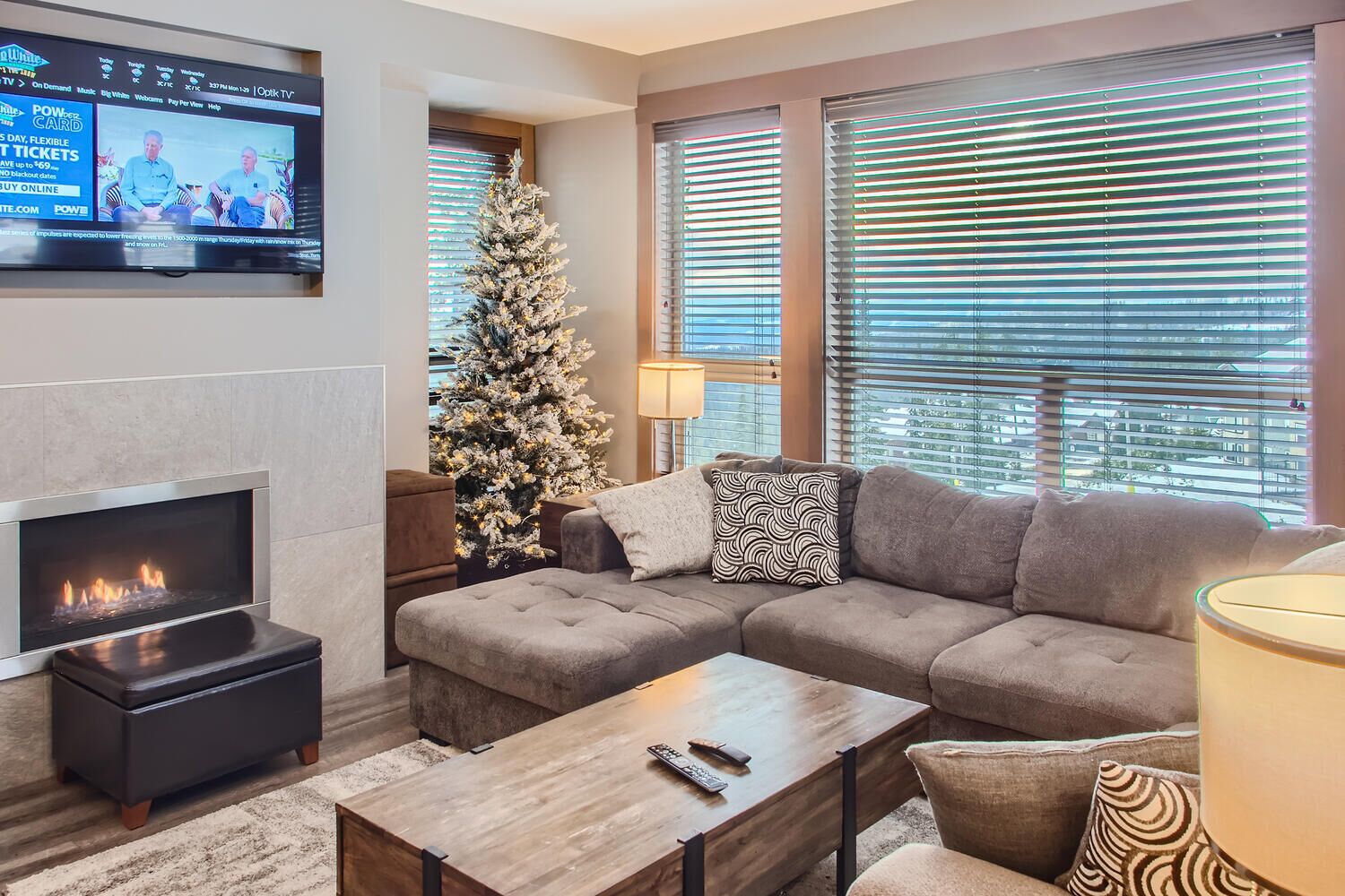 Ein modernes Wohnzimmer mit Kamin, einem großen Fenster mit Jalousien, einem Weihnachtsbaum und einem an der Wand befestigten Fernseher.