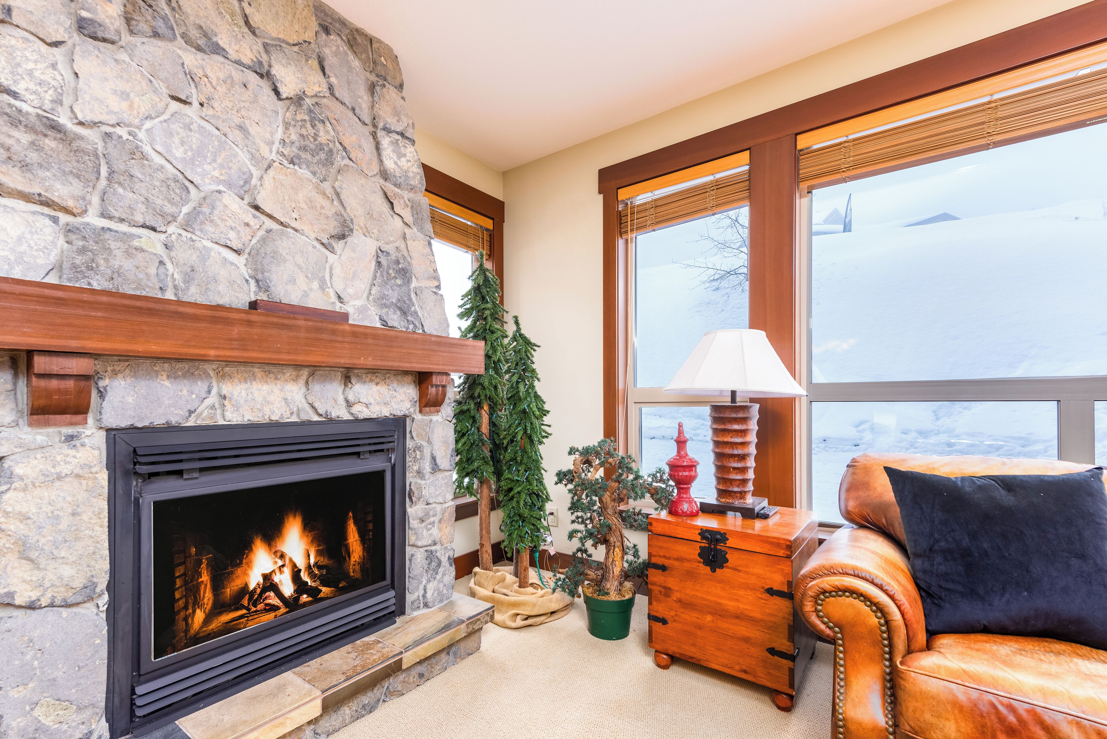 A stone fireplace with a fire, a leather sofa, a wooden chest, and a window with a view of a snowy landscape.