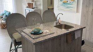 A modern kitchen with a granite countertop, wooden cabinets, and a built-in island with a sink. There are wicker chairs and a dining table in the adjacent living area. - Coral Criff juan dolio (Guayacanes)
