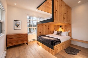 A bedroom with a wooden bunk bed, a bedside table, a dresser, a framed picture, and a window with a view of trees.