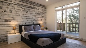 Een slaapkamer met een houten wand, een groot bed, een nachtkastje, een raam met uitzicht op bomen en een balkon.