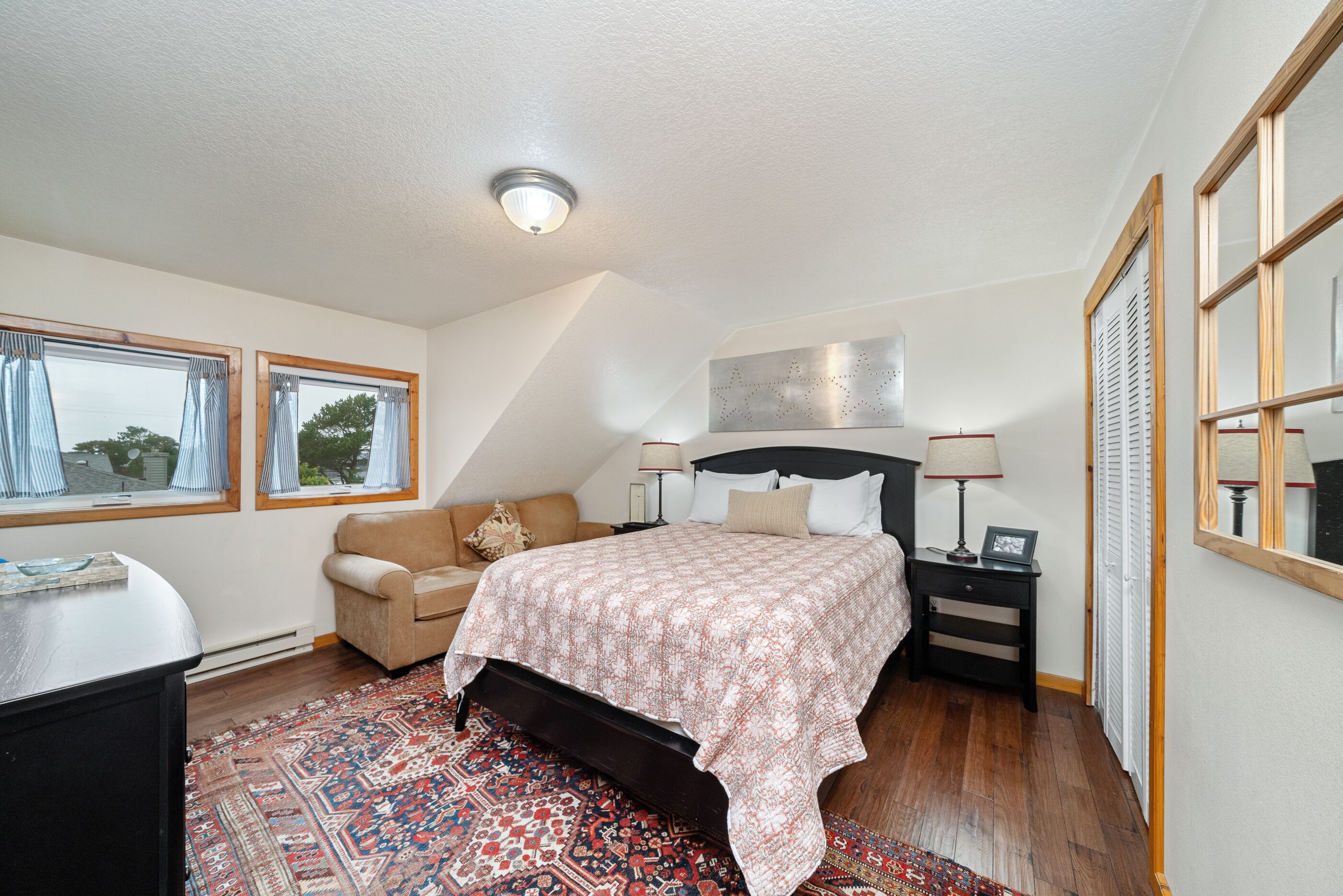 Une chambre à coucher comprenant un lit, une table de chevet, une commode, une fenêtre avec des rideaux, un tapis à motifs et un luminaire au plafond.