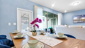 A dining area with a wooden table set for four, a vase of purple flowers, and a view of a living room with a couch and a painting. - Relaxing 5 Bed Townhome with Splash Pool-3966LA (Davenport)
