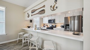 A modern kitchen with white cabinetry, a stainless steel refrigerator, a microwave, and a coffee maker. There are three white bar stools and a large island with a sink. Decorative anchors are mounted on the wall above the island.