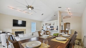 A dining area with a wooden table set for four, a ceiling fan, and a kitchen in the background.