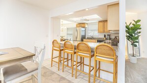 A modern kitchen with white cabinetry, a central island, and a dining area with a wooden table and four wicker chairs. - Beachside Bliss in The Moorings | 3BR Naples Stay (Naples)
