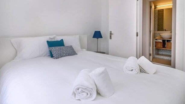 A neatly made bed with white linens and two rolled towels, a blue lamp, and a view of a bathroom with a sink and mirror.