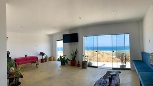 A spacious living room with a large sliding glass door leading to a balcony with a view of the ocean. There are several potted plants and a red sofa in the room. - Deluxe Double Room with Balcony and Sea View (Pis Lobitos)