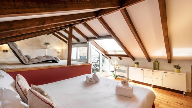 Quarto com teto inclinado, cama com roupa de cama branca, poltrona reclinável, mesinha de cabeceira com abajur e vista pela janela.
