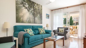 A living room with a teal sofa, a coffee table, and a gray armchair. There is a large window with curtains, a ceiling fan, and a painting of trees on the wall. - Sunset Villa El Olivo (Alcúdia)