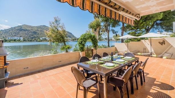 An outdoor dining area with a view of a lake and mountains, equipped with a table set for four, chairs, and umbrellas. - Sunset Villa Isleta (Alcúdia)